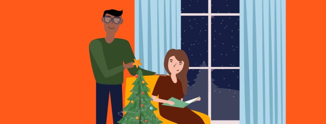 A man puts a star atop a Christmas tree and stands next to a woman sitting in a chair with a book